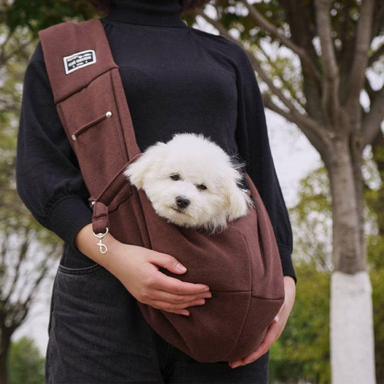 Bolsa de ombro marrom para pets, com um cachorro branco dentro, mostrada sendo segurada por uma pessoa.